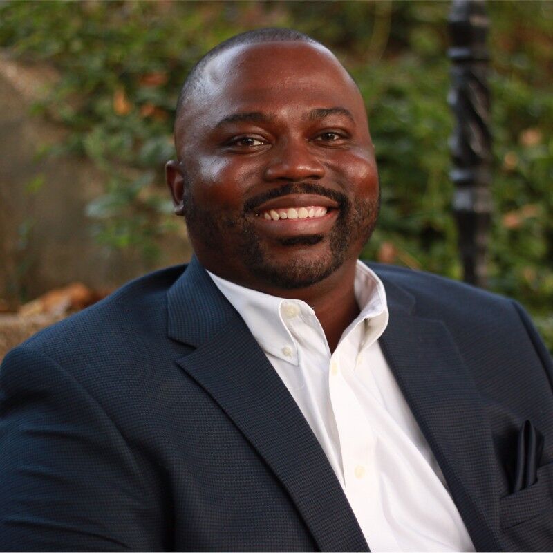 The image shows a smiling Black man in a navy blue suit jacket and a white shirt. He is sitting outside with greenery in the background. He appears to be in his late 30s or early 40s. The man has a neatly trimmed beard and short hair.